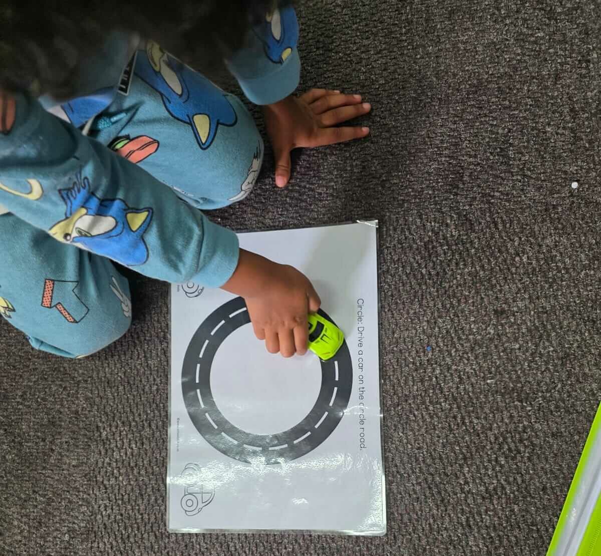 Pre-writing tracing activity at Petra Christian Pre-School — a young child in blue pyjama-themed clothing traces a circle road shape on a laminated activity sheet using a small toy car