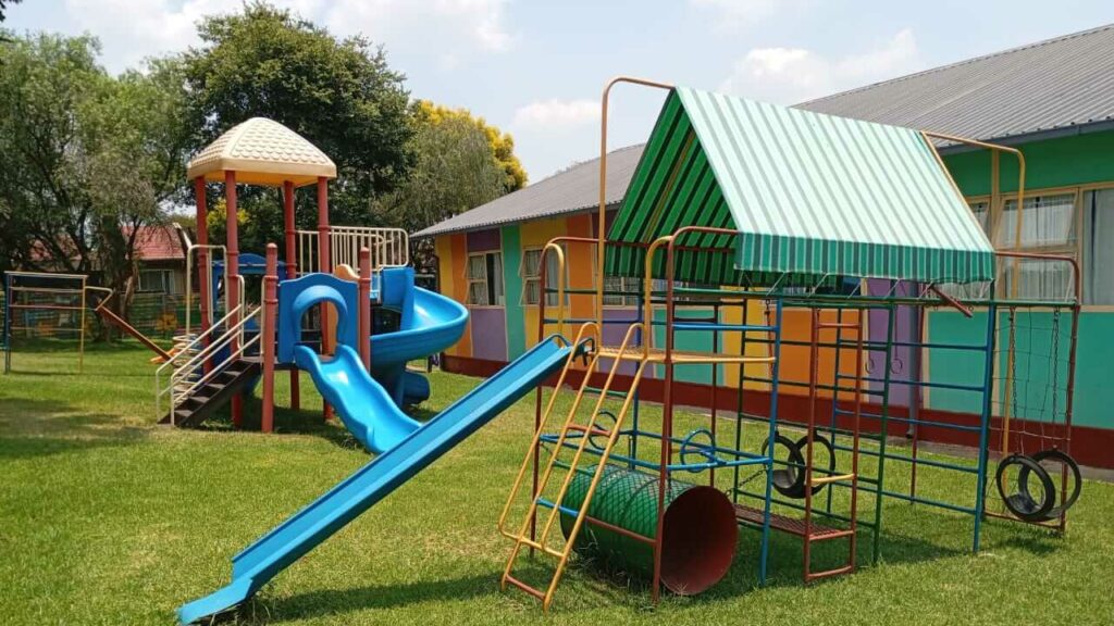Alt Text Outdoor playground at Petra Christian Pre-School, Edenvale, Johannesburg — featuring colourful jungle gyms, a blue slide, climbing frames with tyres and a crawl tunnel, a shaded play structure with shaded canopy