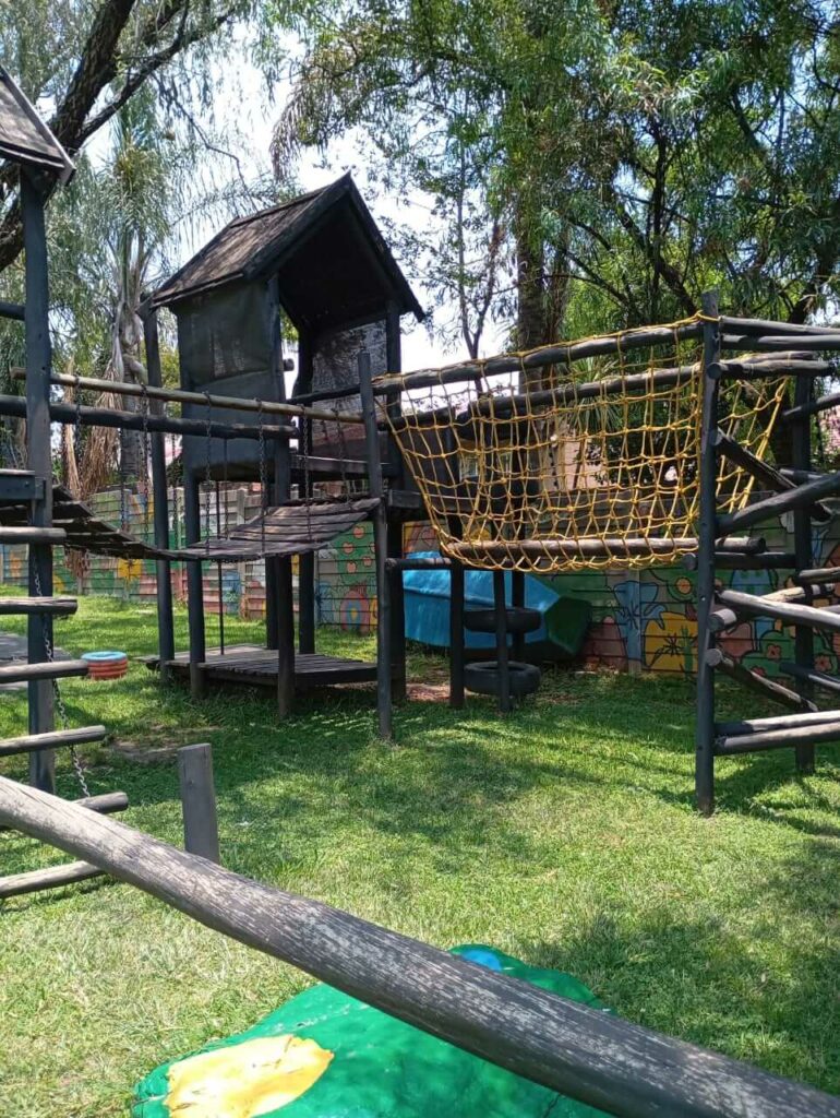 Spacious garden and outdoor play area at Petra Christian Pre-School, Edenvale — shaded by mature trees with lush green lawns, multiple play structures, pathways, and a colourful hand-painted animal mural on the fully enclosed perimeter wall.