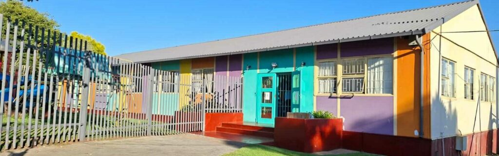Petra Christian Pre-School building exterior in Marais Steyn Park, Edenvale, Johannesburg — a vibrant, colourfully painted single-storey school with teal, purple, orange, and yellow panels, a secure gated entrance, lush green lawn, and a clear blue sky.