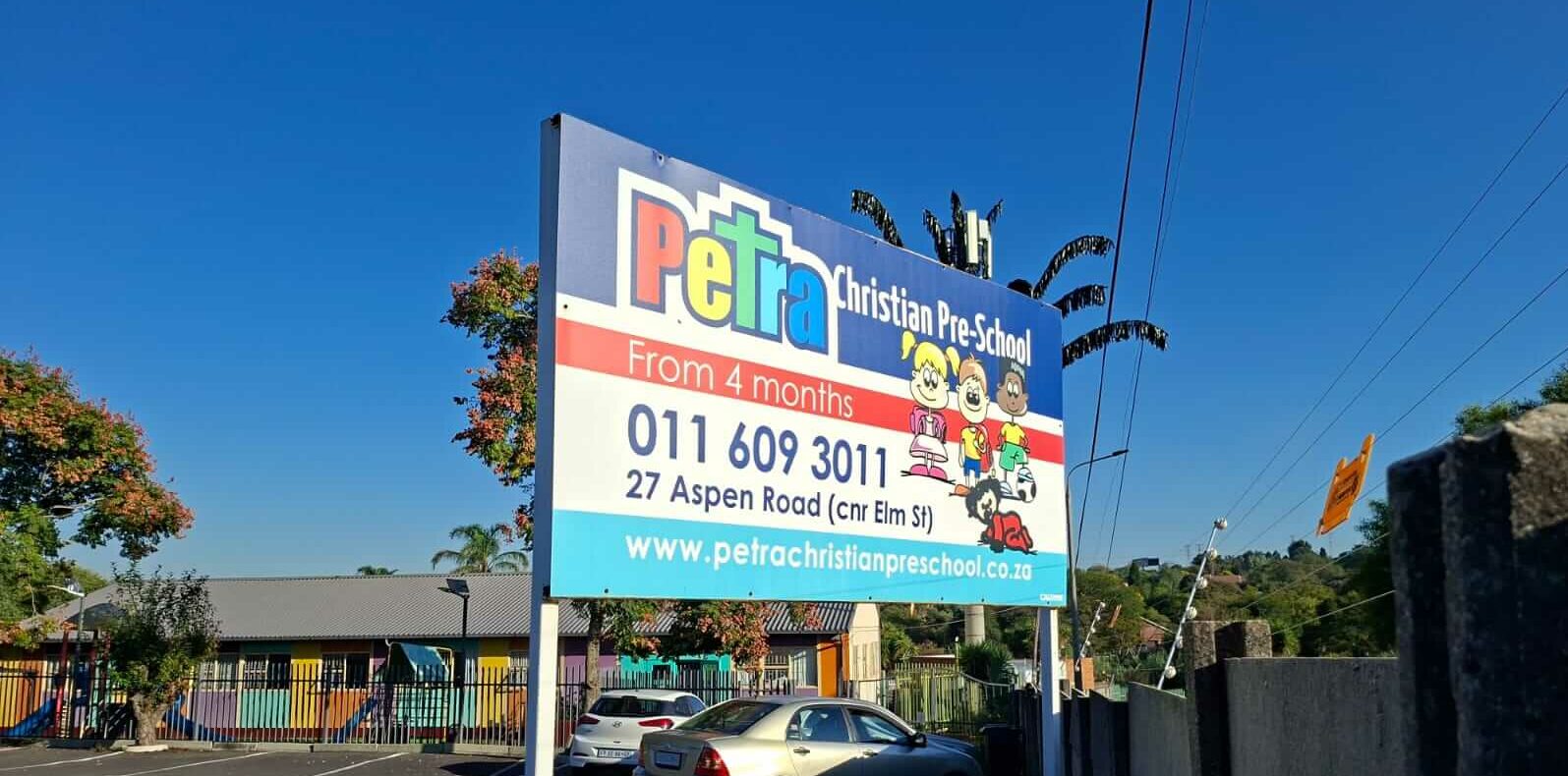 Street-facing signage board for Petra Christian Pre-School, Edenvale — colourful sign reading "Petra Christian Pre-School, From 4 months, 011 609 3011, 27 Aspen Road (cnr Elm St) Marais Steyn Park